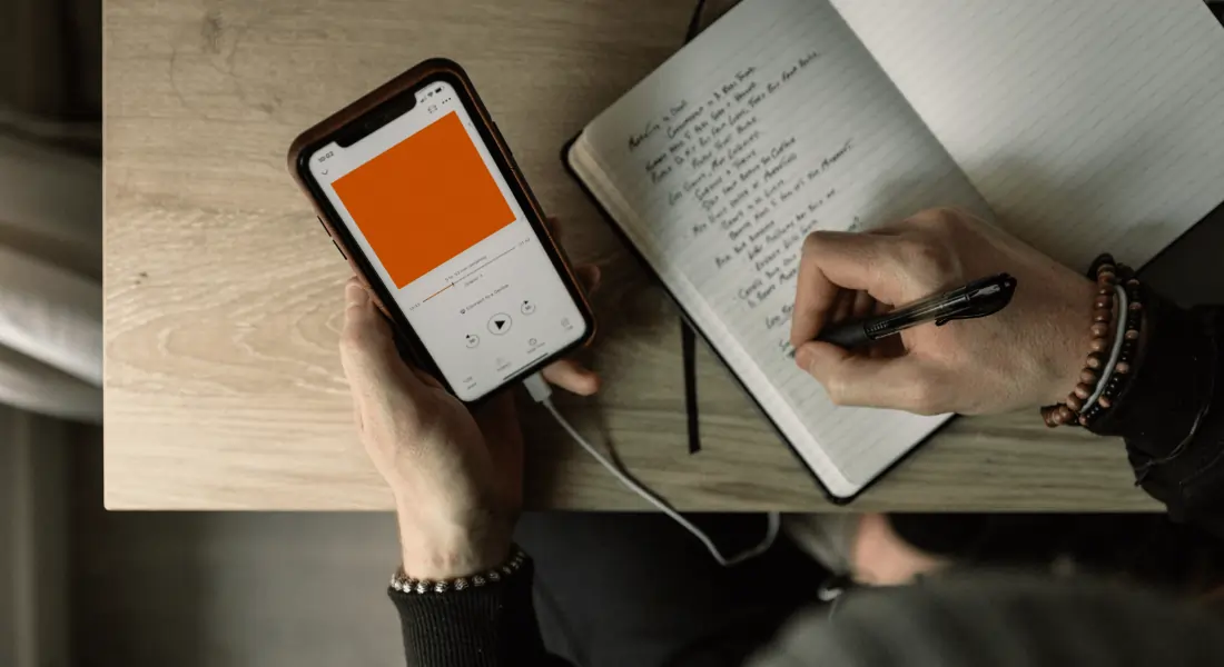 A person listening to a Native Mobile App (possibly a podcast) while making notes simultaneously using a black pen.